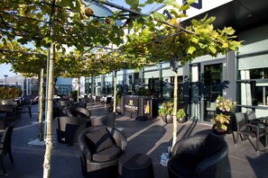 Un espacio al aire libre con mobiliario de mimbre, una zona de bar y un edificio con grandes ventanales.