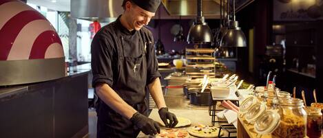 A chef preparing pizzas in a commercial kitchen with stainless steel surfaces, overhead lighting, and various food containers.