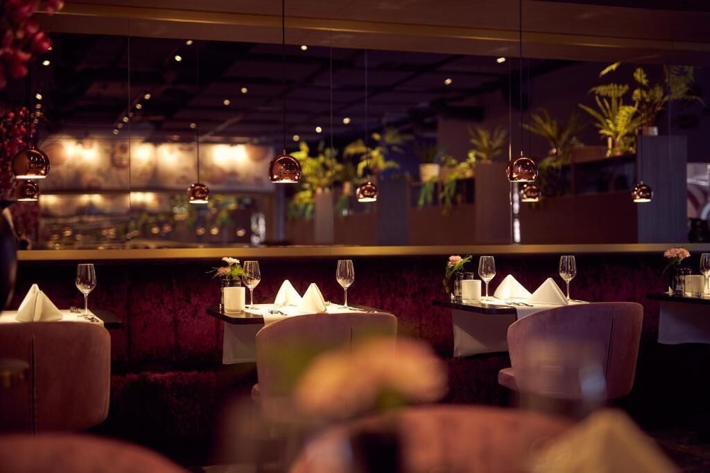 Un interior de restaurante con mesas dispuestas para comer, lámparas colgantes y plantas en macetas al fondo.