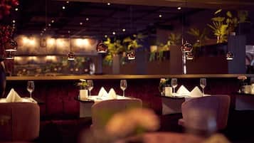 Un interior de restaurante con mesas dispuestas para comer, lámparas colgantes y plantas en macetas al fondo.