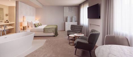A modern hotel room with a large bathtub, a bed with green bedding, a seating area with two chairs, a small round table, and a flat-screen TV mounted on the wall.