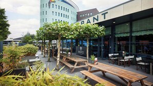 An outdoor dining area with wooden benches and tables, surrounded by greenery and a building with a sign that reads "ROOTZ". - Van Der Valk Hotel Amersfoort A1 (Amersfoort)