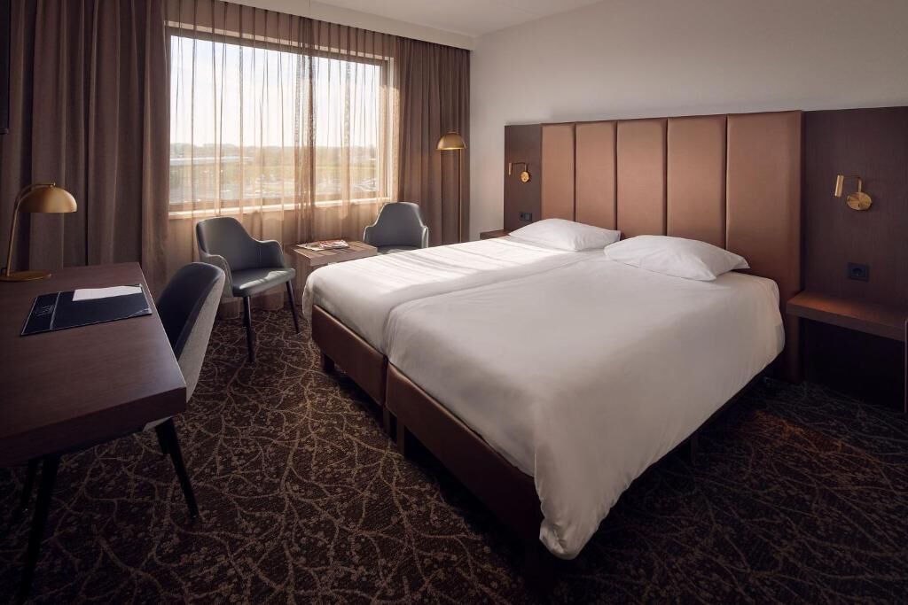 Quarto de hotel com uma cama grande, uma escrivaninha, duas cadeiras, uma mesinha e uma janela com cortinas.