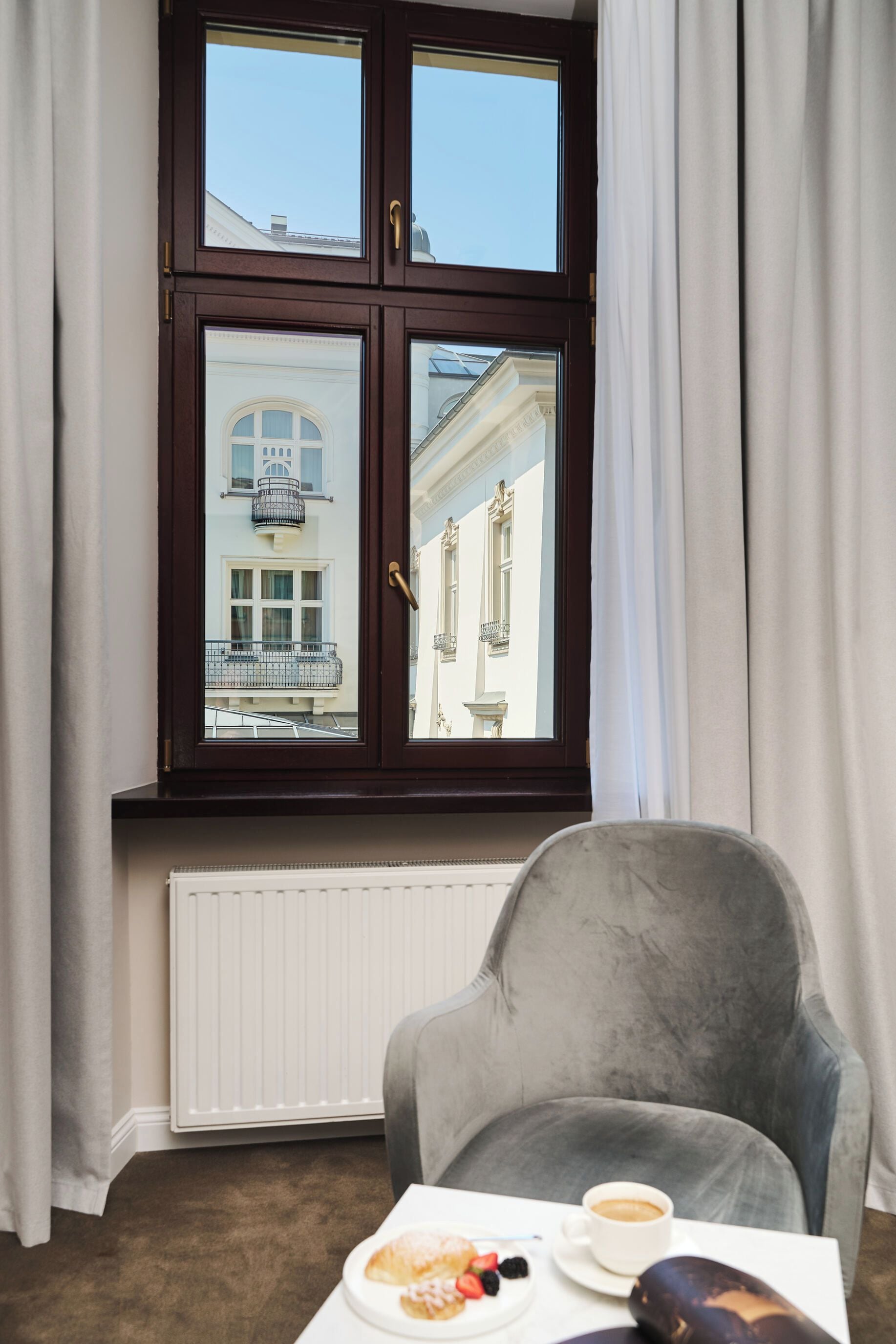 A room with a window, a radiator, a grey armchair, a table with a plate of food and a cup of coffee.