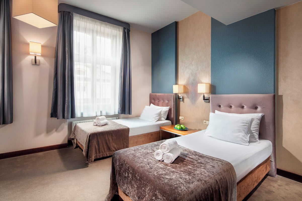 A hotel room with two beds, a large window with curtains, wall-mounted lamps, and a small table with a fruit bowl.