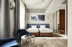 A hotel room with two beds, a blue chair, a small table with a tray of items, and a large window with curtains.