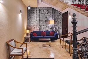 Um hall com sofá azul, cadeiras de madeira, mesa de centro, parede estampada e escada com degraus vermelhos.