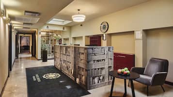 A hotel lobby with a reception desk, a clock, a bookshelf, and a seating area with a table and chairs.
