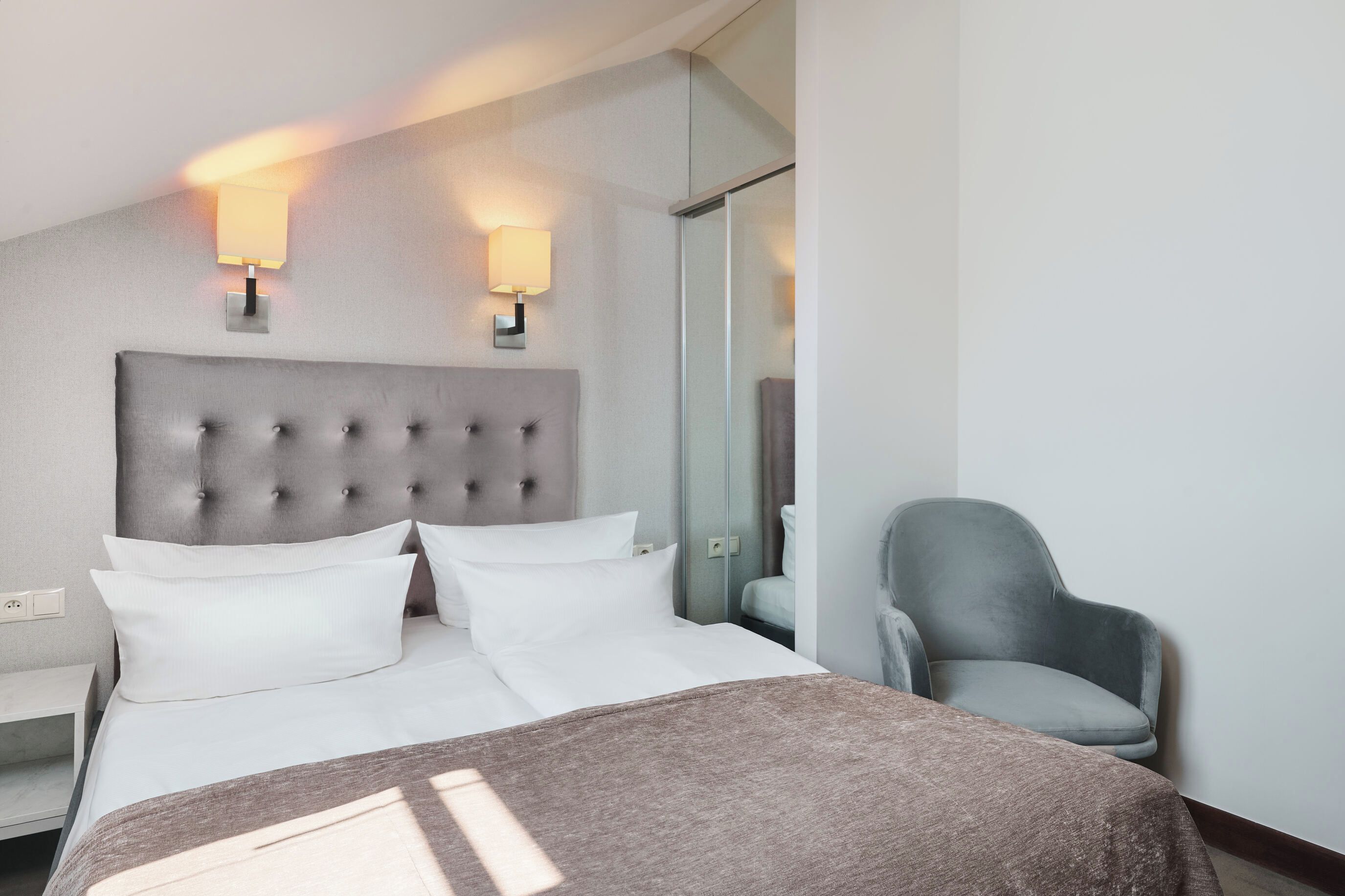 A hotel room with a large bed, two wall-mounted lamps, a grey armchair, and a bedside table.