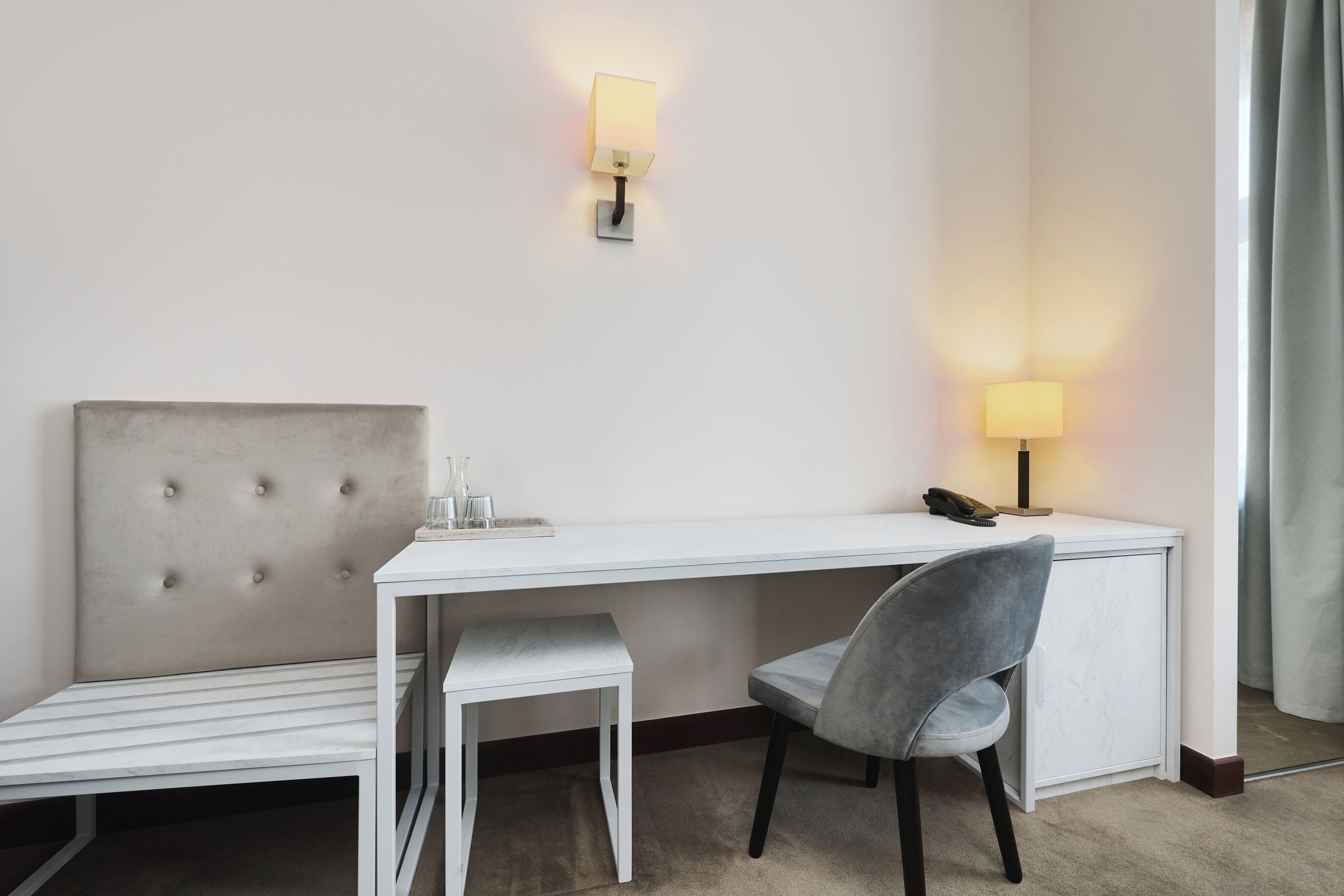 A hotel room with a white desk, a grey upholstered chair, a bedside table, and a lamp.