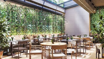 An indoor dining area with tables and chairs, surrounded by lush green plants and a high ceiling with skylights.