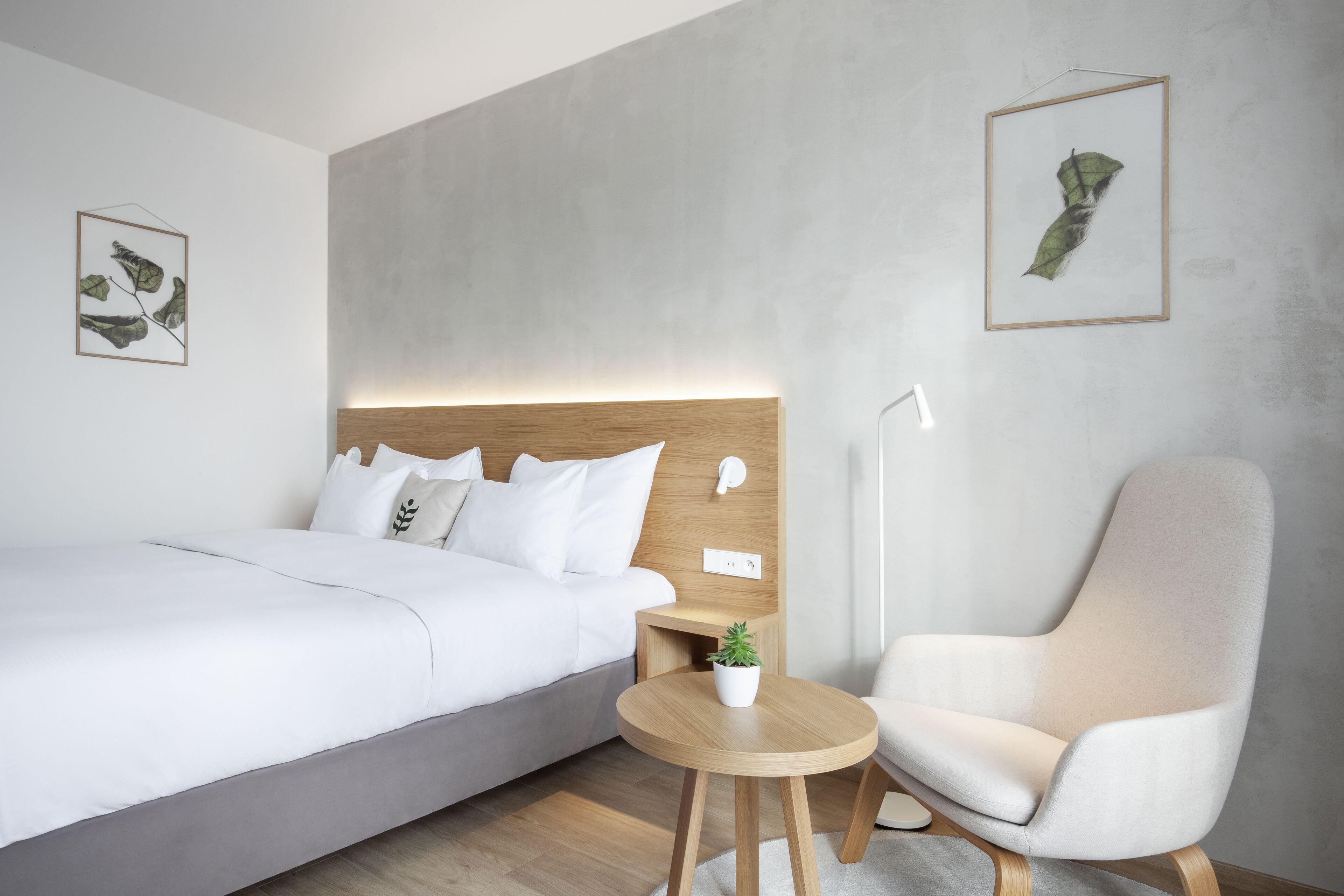 A modern hotel room with a bed, a chair, a small table with a plant, and artwork on the walls.