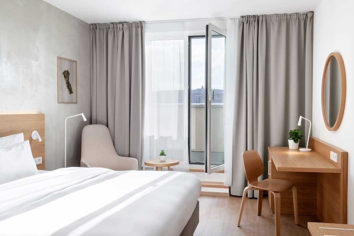 A hotel room with a bed, a desk, a chair, a small table with a plant, and a view through a glass door.