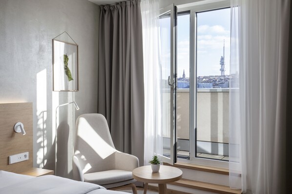 Une chambre d’hôtel moderne dotée d’une grande porte-fenêtre coulissante donnant sur un balcon, d’un fauteuil confortable, d’une petite table avec une plante et d’un décor minimaliste.