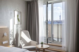 Quarto de hotel moderno com uma grande porta de vidro deslizante para uma varanda, uma poltrona confortável, uma pequena mesa com uma planta e decoração minimalista.