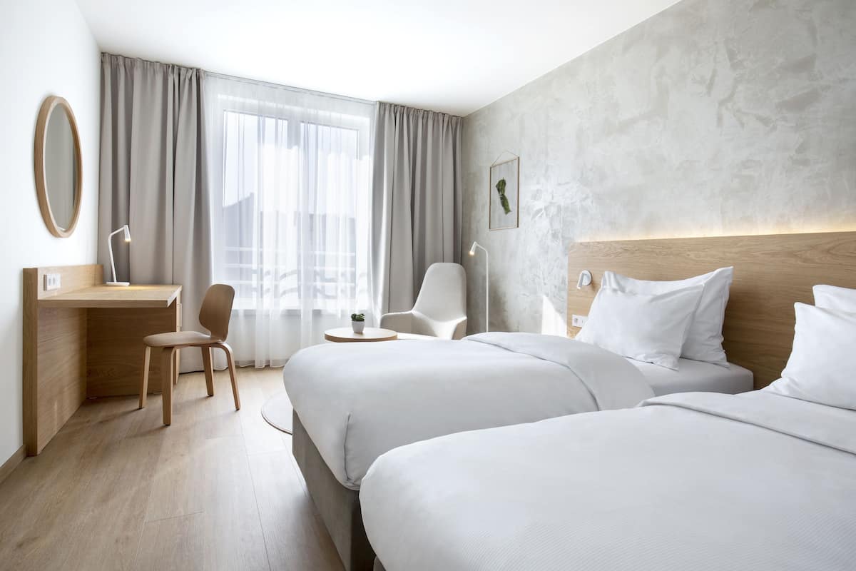 A hotel room with two beds, a desk, a chair, and a large window with curtains.