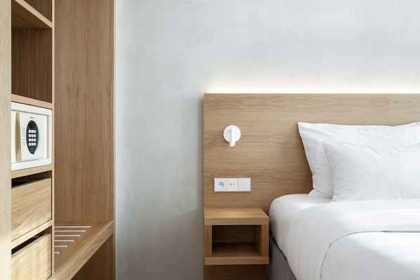 A neatly organized hotel room with a wooden headboard, a bedside table with a lamp, and a built-in safe.