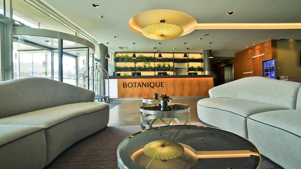A modern lobby with a reception desk labeled "BOTANIQUE," featuring a glass partition, comfortable seating, and a unique round table with a central light fixture. - Botanique Hotel Prague (Prague)