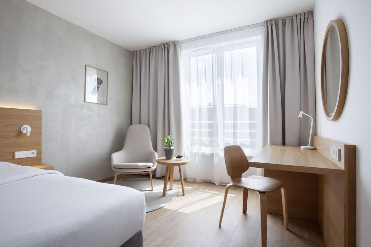 A modern hotel room with a bed, a desk, two chairs, a small table with a plant, and a large window with curtains.