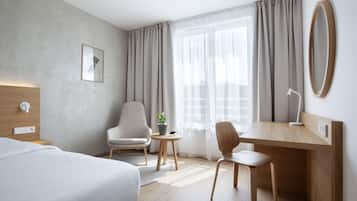Quarto de hotel moderno com cama, escrivaninha, duas cadeiras, uma mesinha de centro com planta e uma janela grande com cortinas.