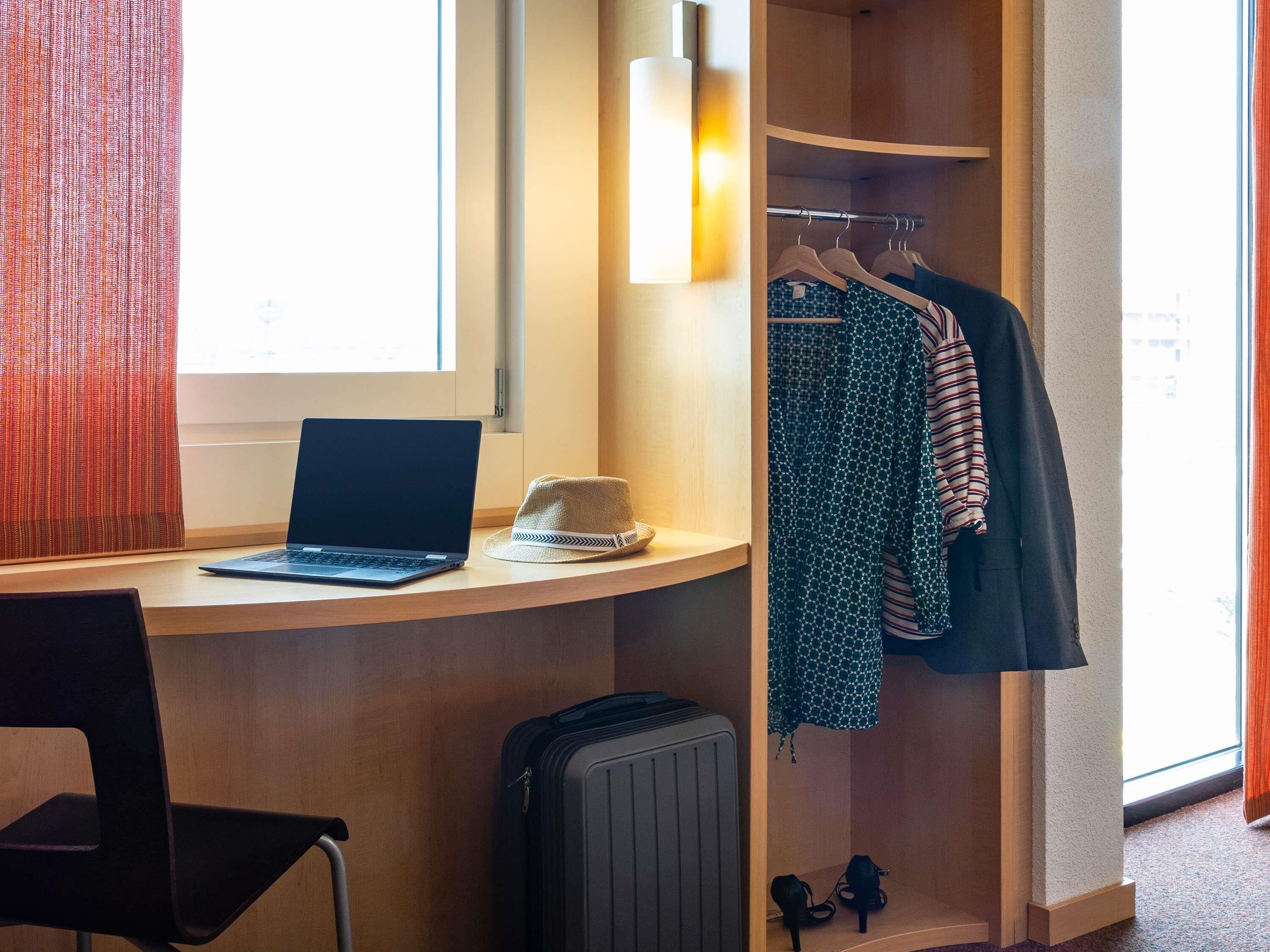 Une chambre d’hôtel équipée d’un bureau, d’un ordinateur portable, d’une valise et d’un portant à vêtements sur lequel sont disposés différents vêtements.