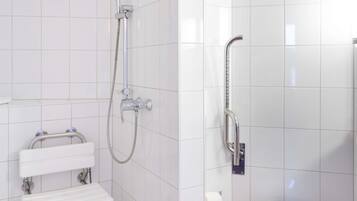 Une salle de bain avec des murs carrelés de blanc, une douche équipée d’un pommeau de douche à main, des toilettes dotées d’une barre d’appui et un lavabo blanc avec un porte-serviettes.