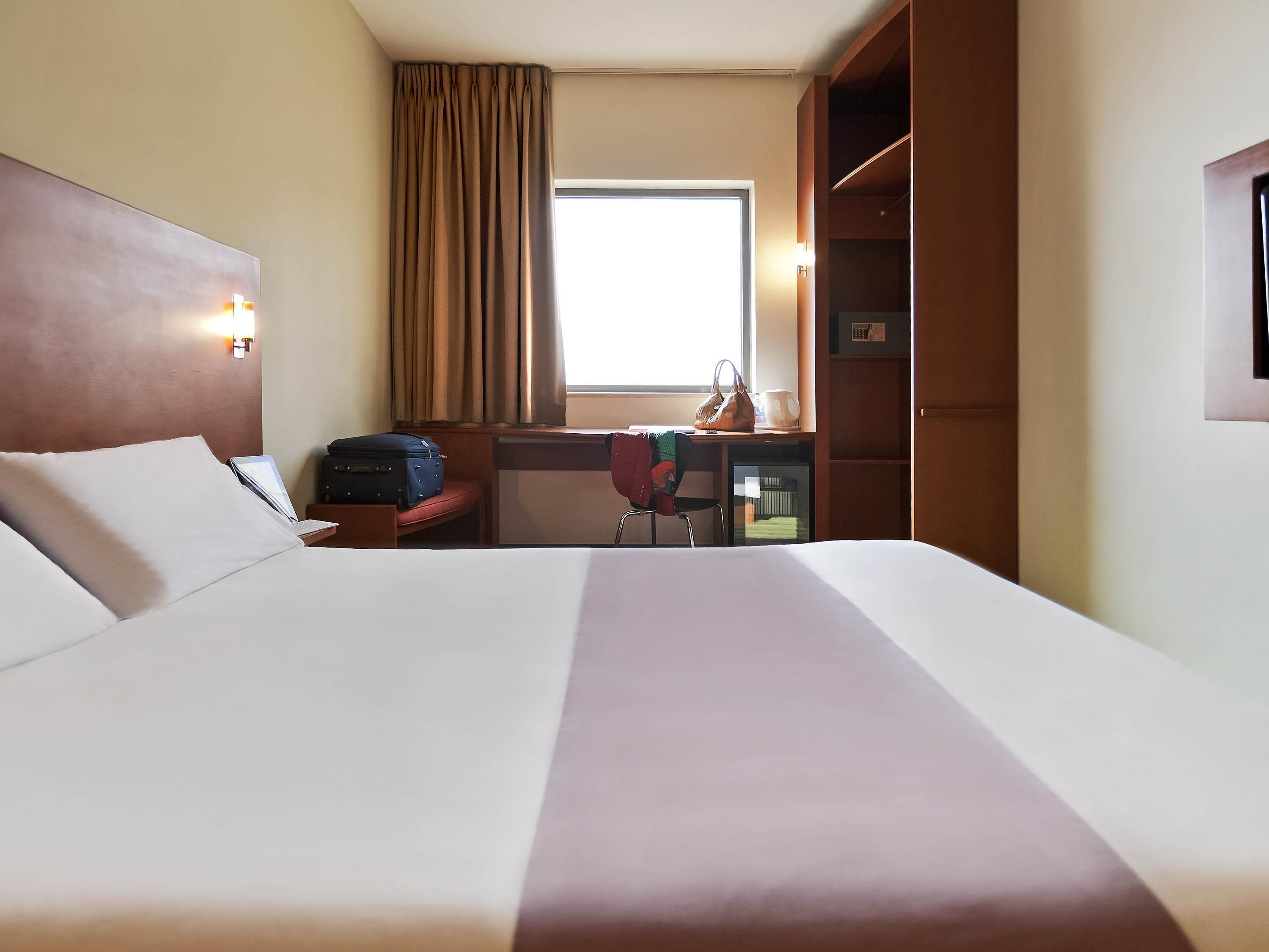 A hotel room with a large bed, a desk with a chair, a suitcase, a handbag, and a window with curtains.