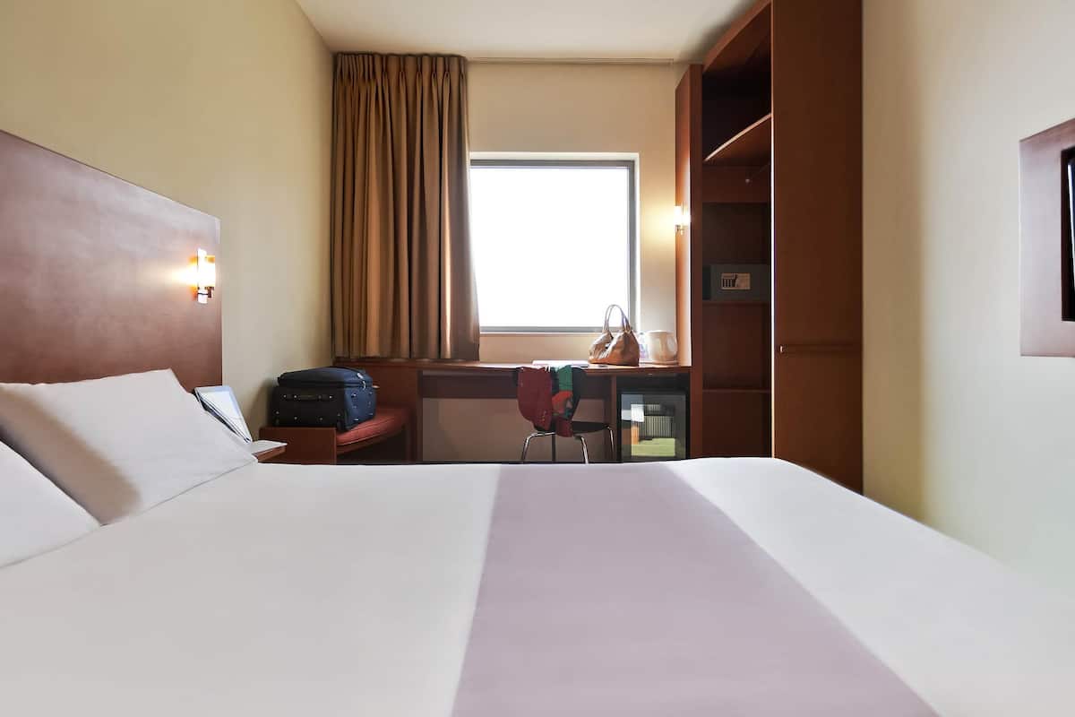 Habitación de hotel con una cama grande, un escritorio con silla, una maleta, un bolso de mano y una ventana con cortinas.