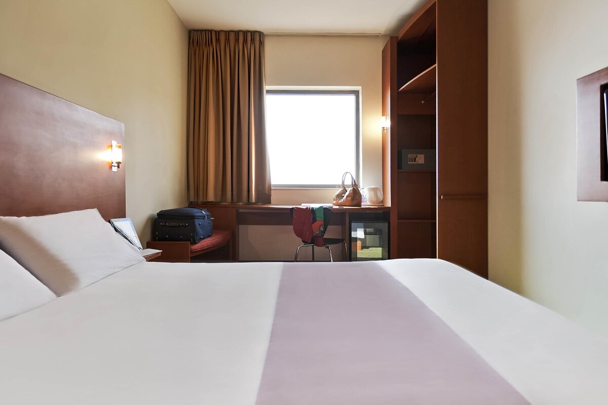 A hotel room with a large bed, a desk with a chair, a suitcase, a handbag, and a window with curtains.