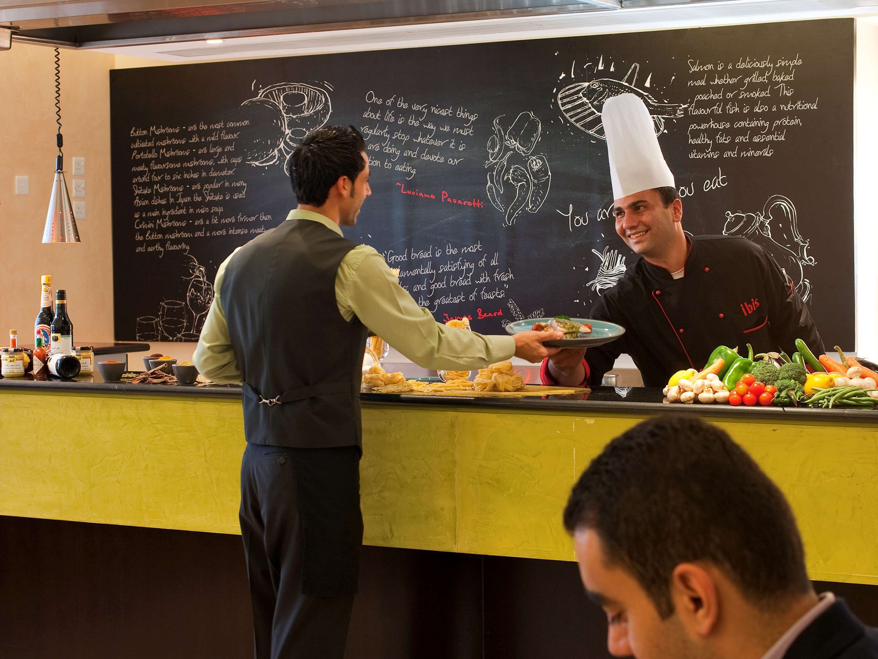Eine Restaurantbedienung mit Koch und Kellner. Die Theke weist eine Schiefertafel mit Illustrationen und Beschreibungen von Speisen auf. Es liegen verschiedene Speisen und Gewürze auf der Theke.