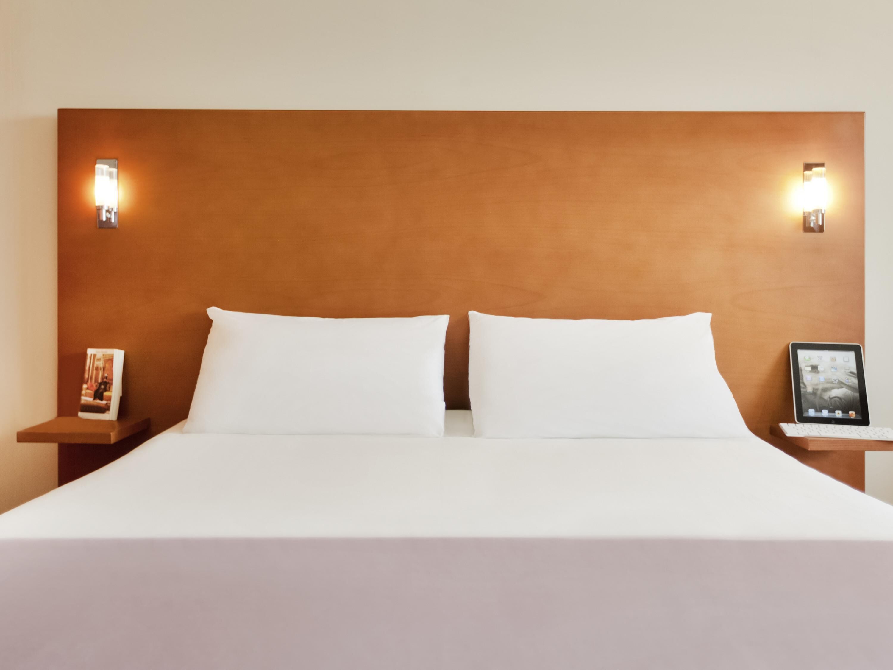 A bed with white linens, two pillows, a wooden headboard, and two bedside tables with wall-mounted lamps.