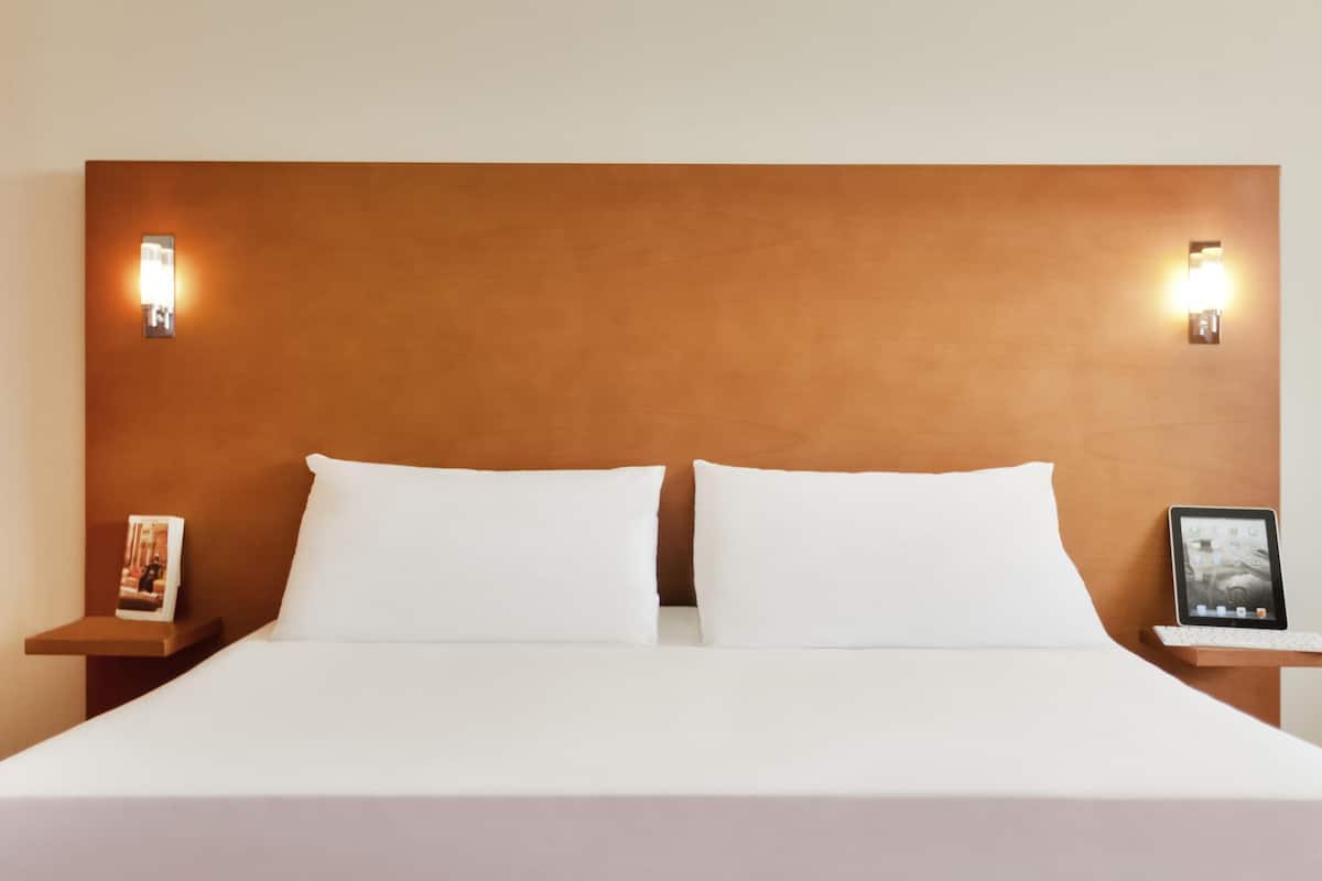 Una cama con sábanas blancas, dos almohadas, un cabecero de madera y dos mesitas de noche con lámparas de pared.