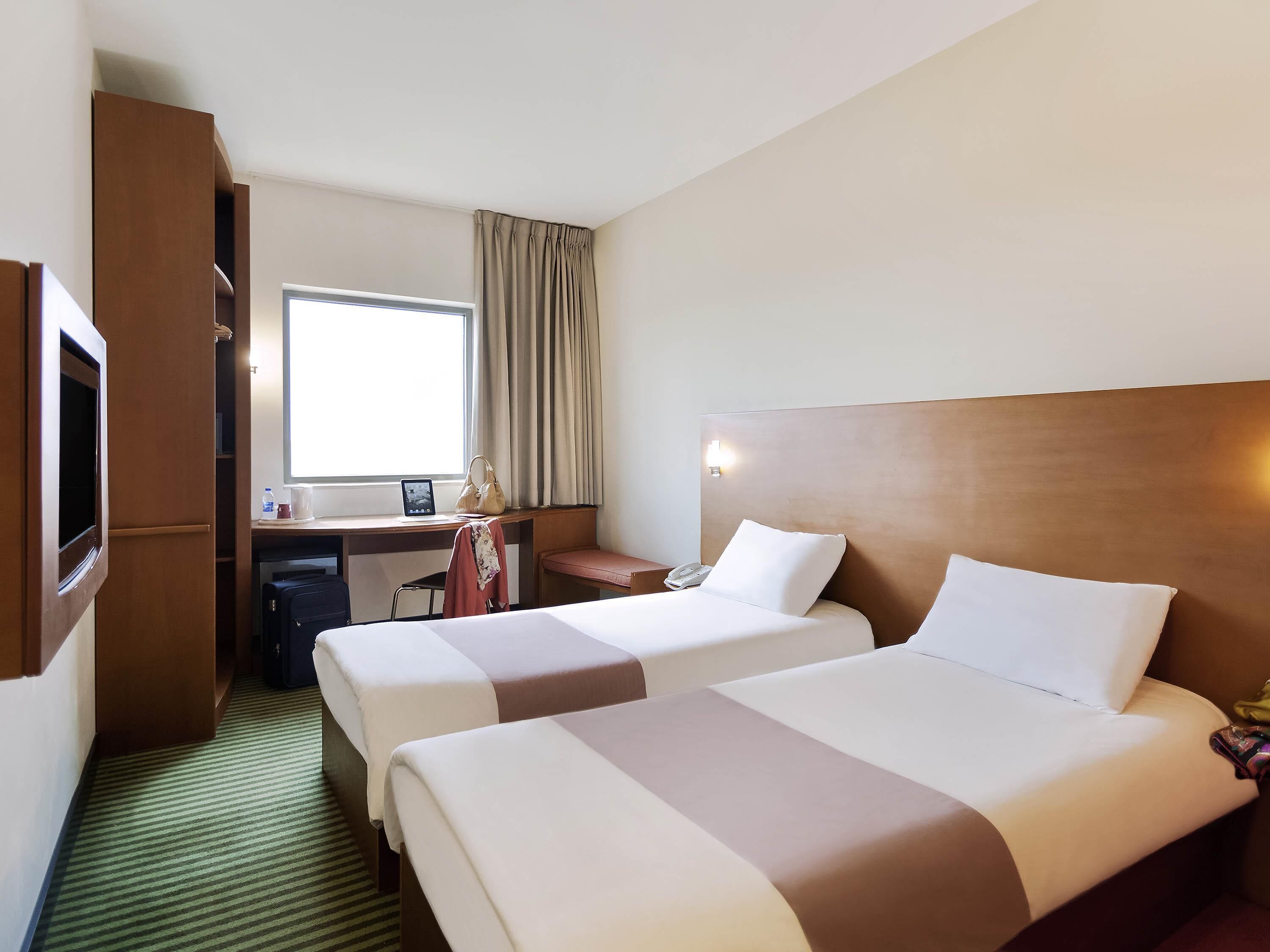 A hotel room with two beds, a desk, a chair, a wardrobe, and a window with curtains.