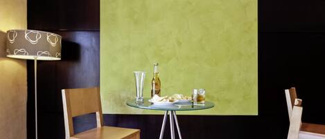 Une pièce avec une table en verre mise pour deux personnes, une bouteille et un verre posés sur la table. Il y a deux chaises et une lampe.