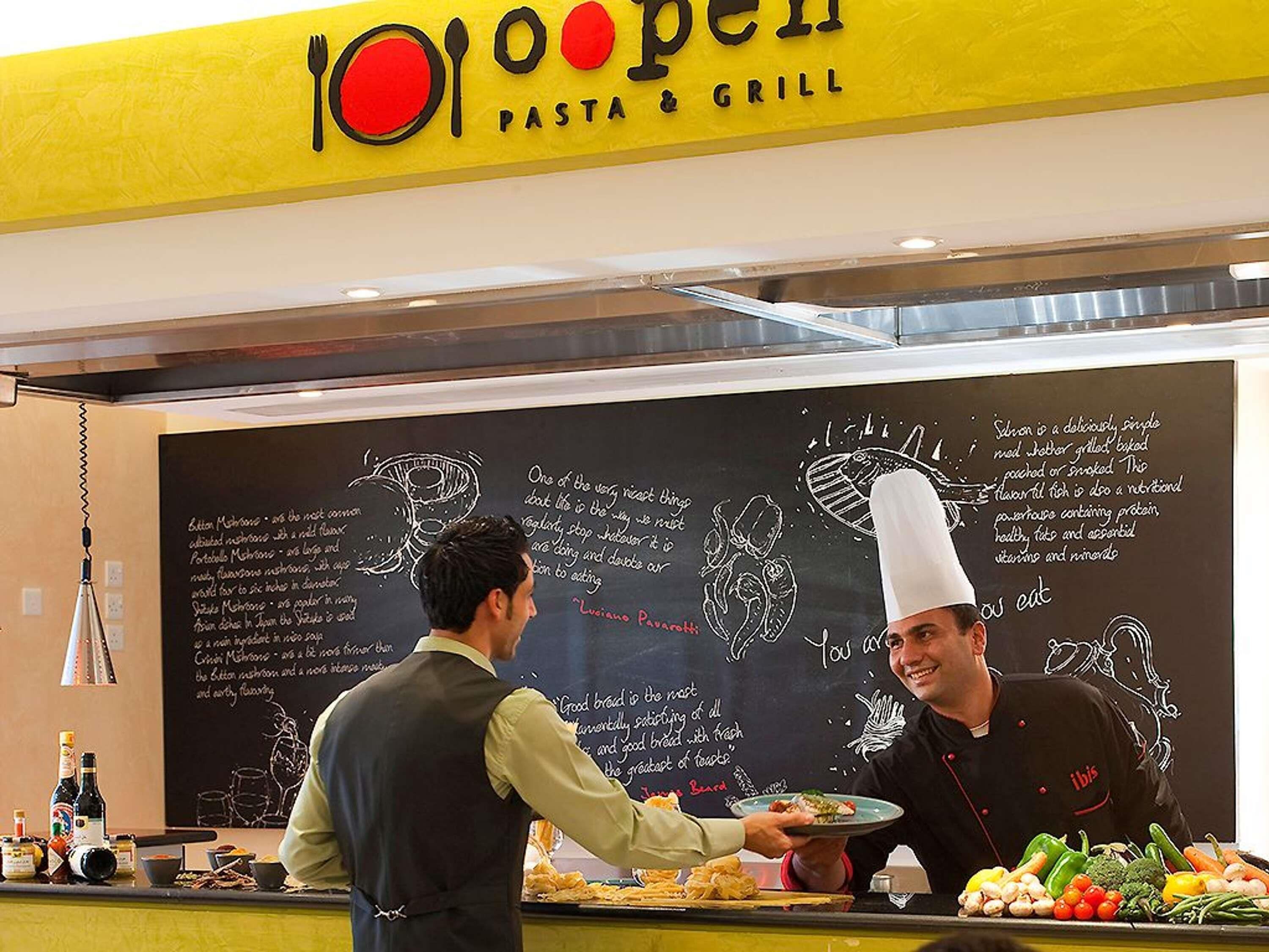 A restaurant interior with a chalkboard menu, a chef serving food, and a variety of fresh vegetables on the counter.