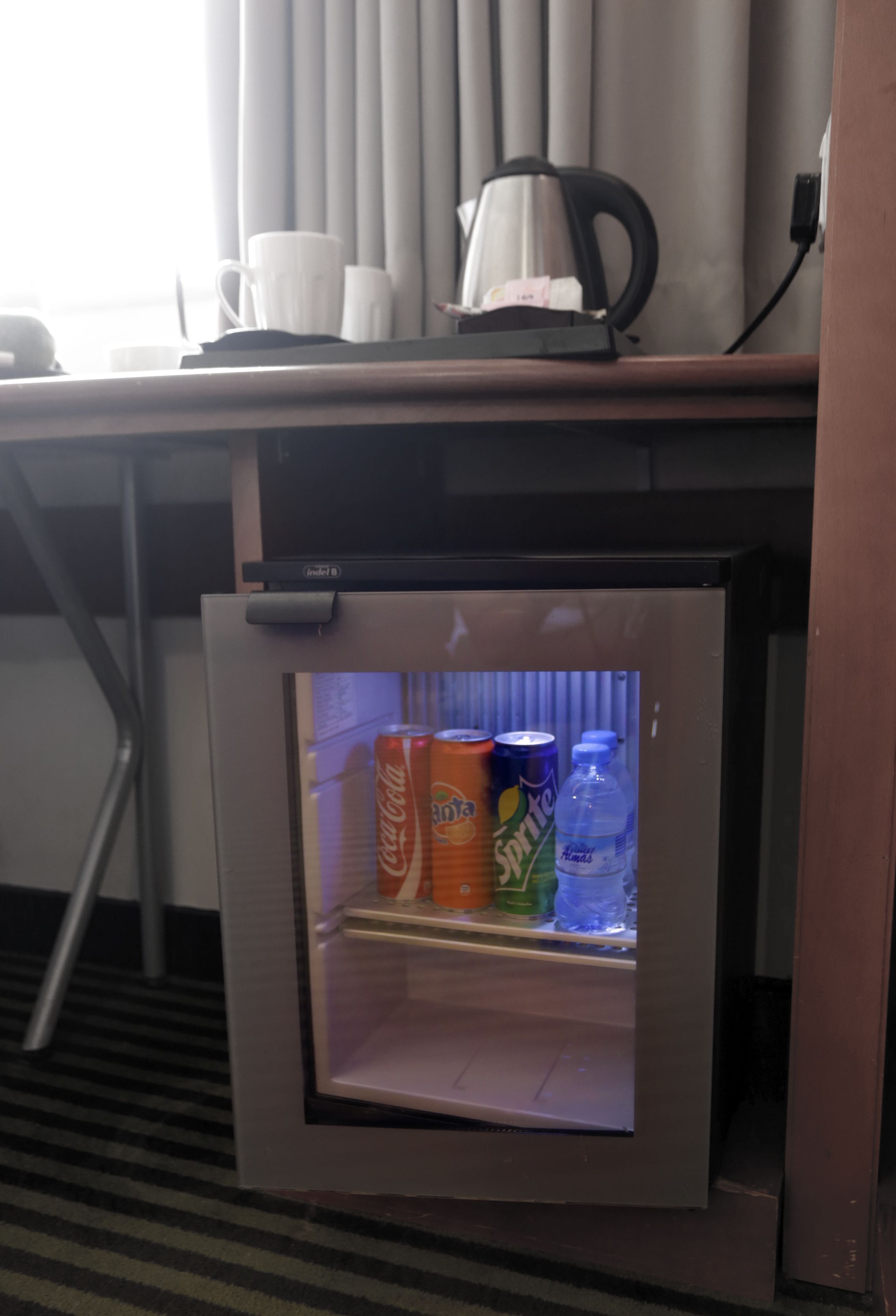 Un refrigerador compacto con puerta de vidrio, que contiene algunas latas de refresco y una botella de agua.