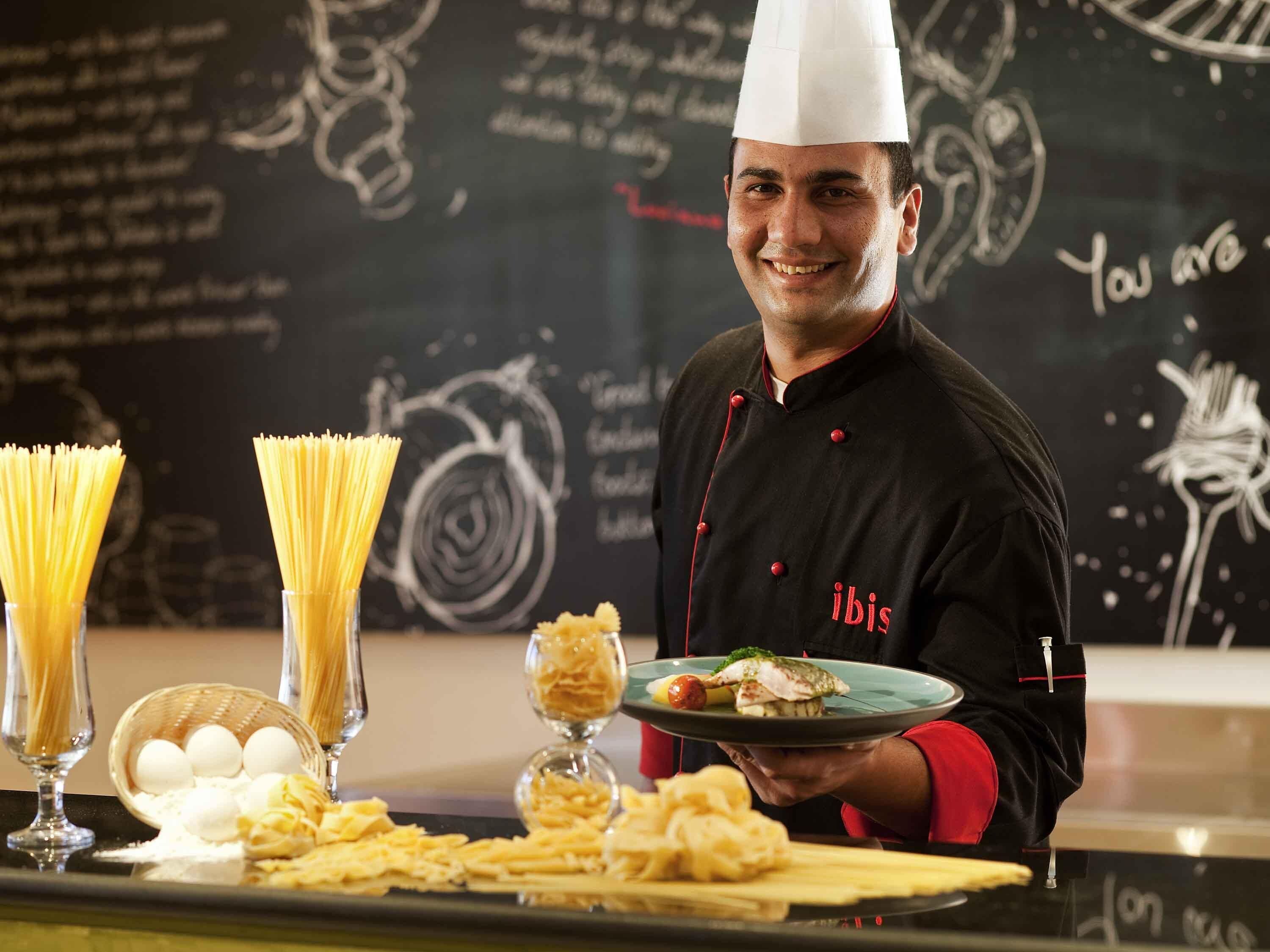 A chef in a black and red uniform holding a plate with food, standing in front of a chalkboard with writing.