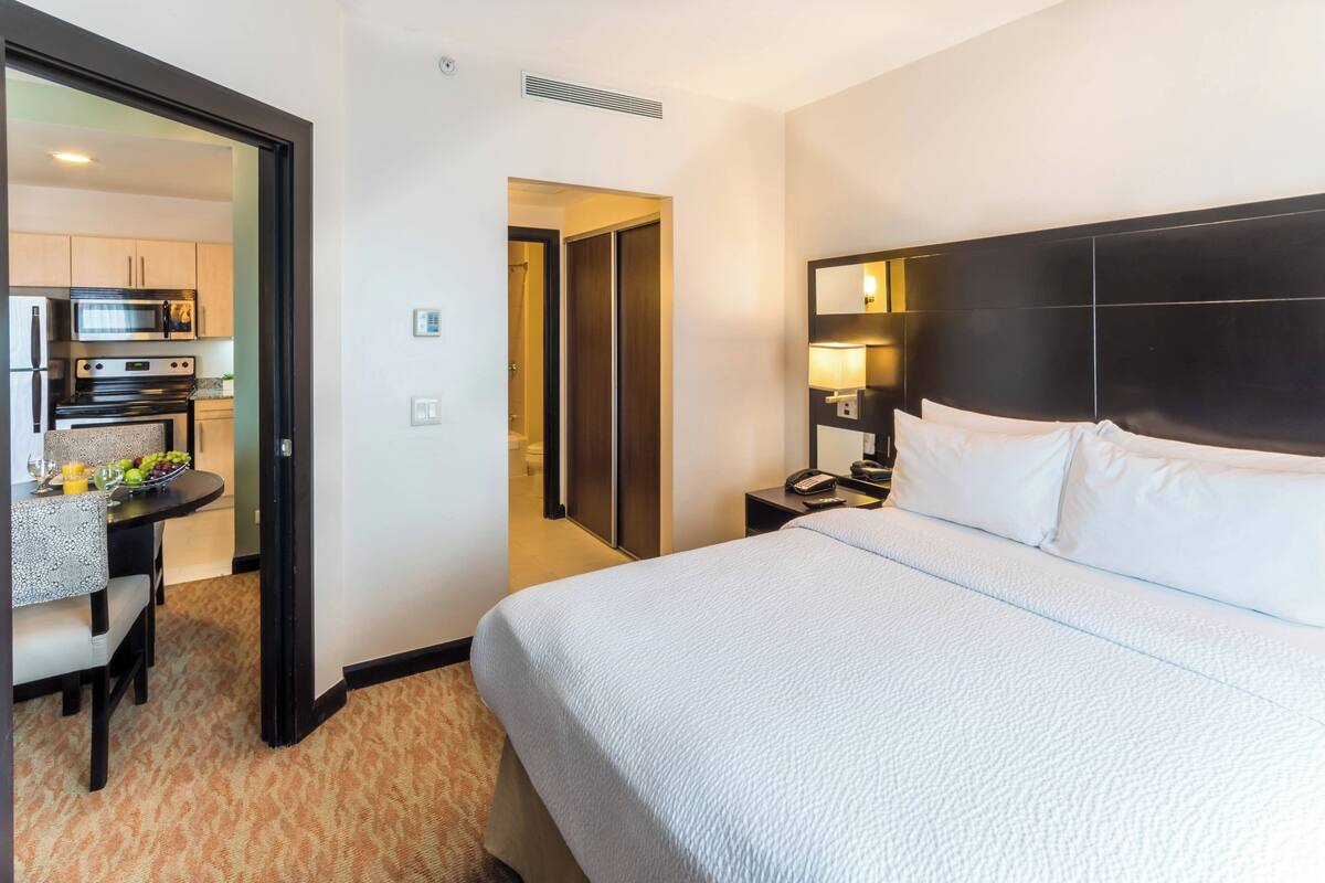 A hotel room with a large bed, a bedside table with a lamp, a small desk with a chair, and a kitchenette in the background.