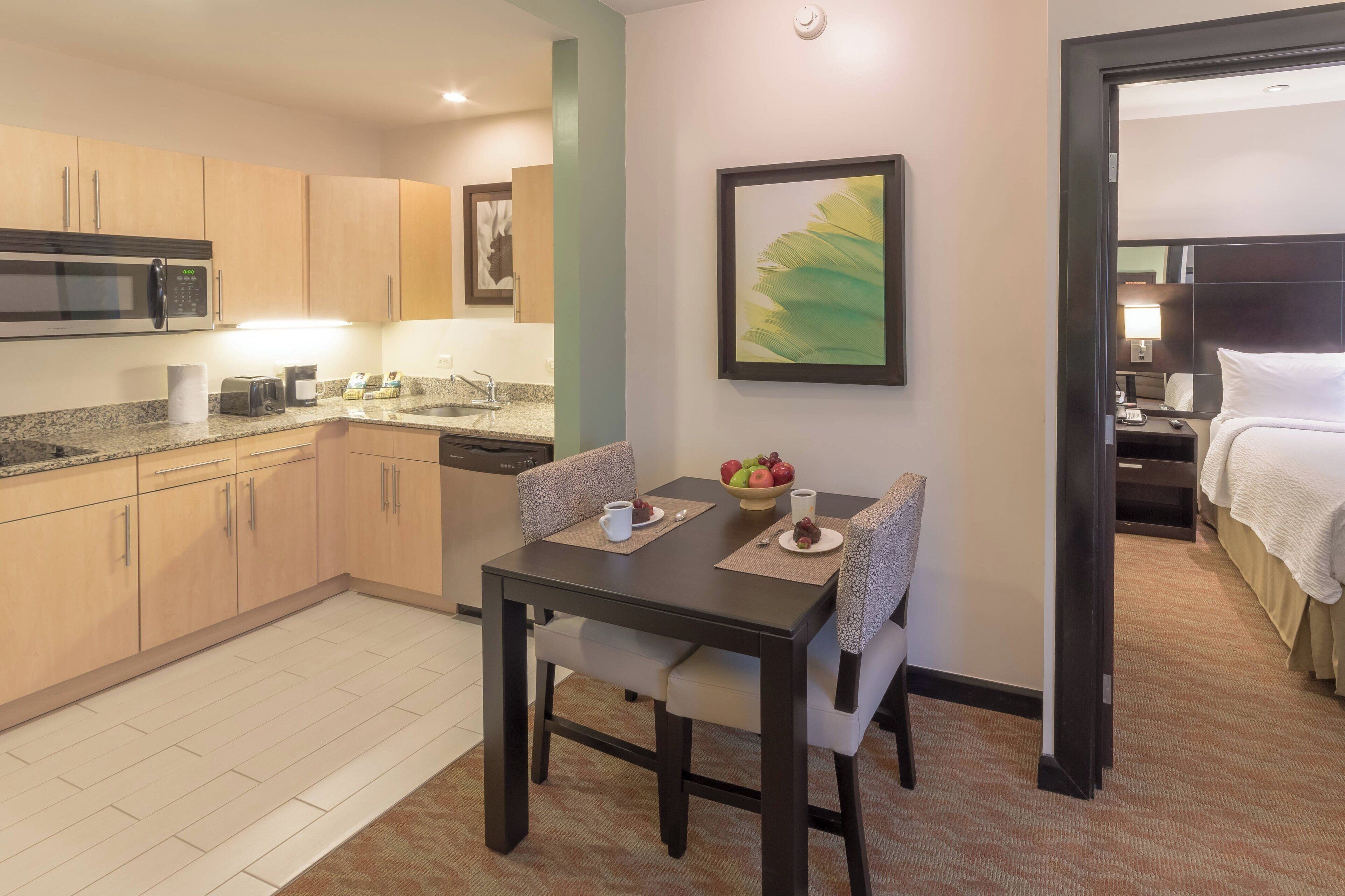 A hotel room with a kitchenette, a dining area with a table and chairs, a bed with a white comforter, and a painting on the wall.