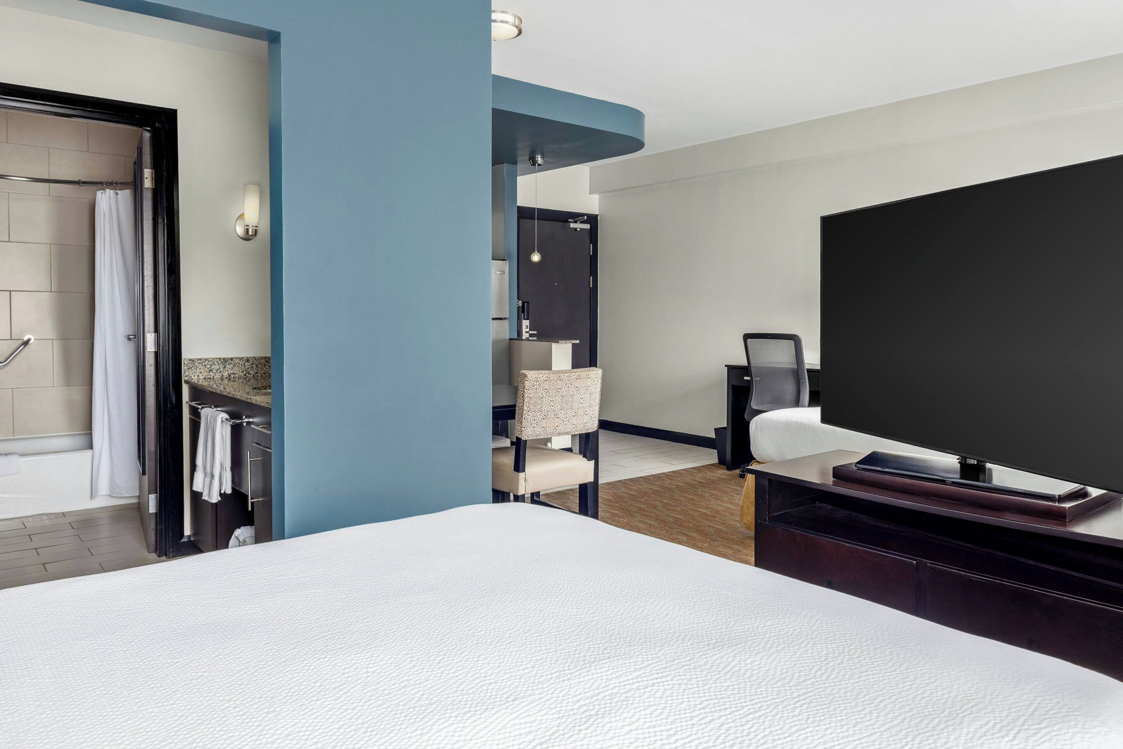 Hotelkamer met een groot bed, een flatscreen televisie, een zithoek met een stoel en een klein tafeltje, een badkamer met douche en handdoekrek, en een blauwe accentmuur.