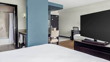 Hotelkamer met een groot bed, een flatscreen televisie, een zithoek met een stoel en een klein tafeltje, een badkamer met douche en handdoekrek, en een blauwe accentmuur.