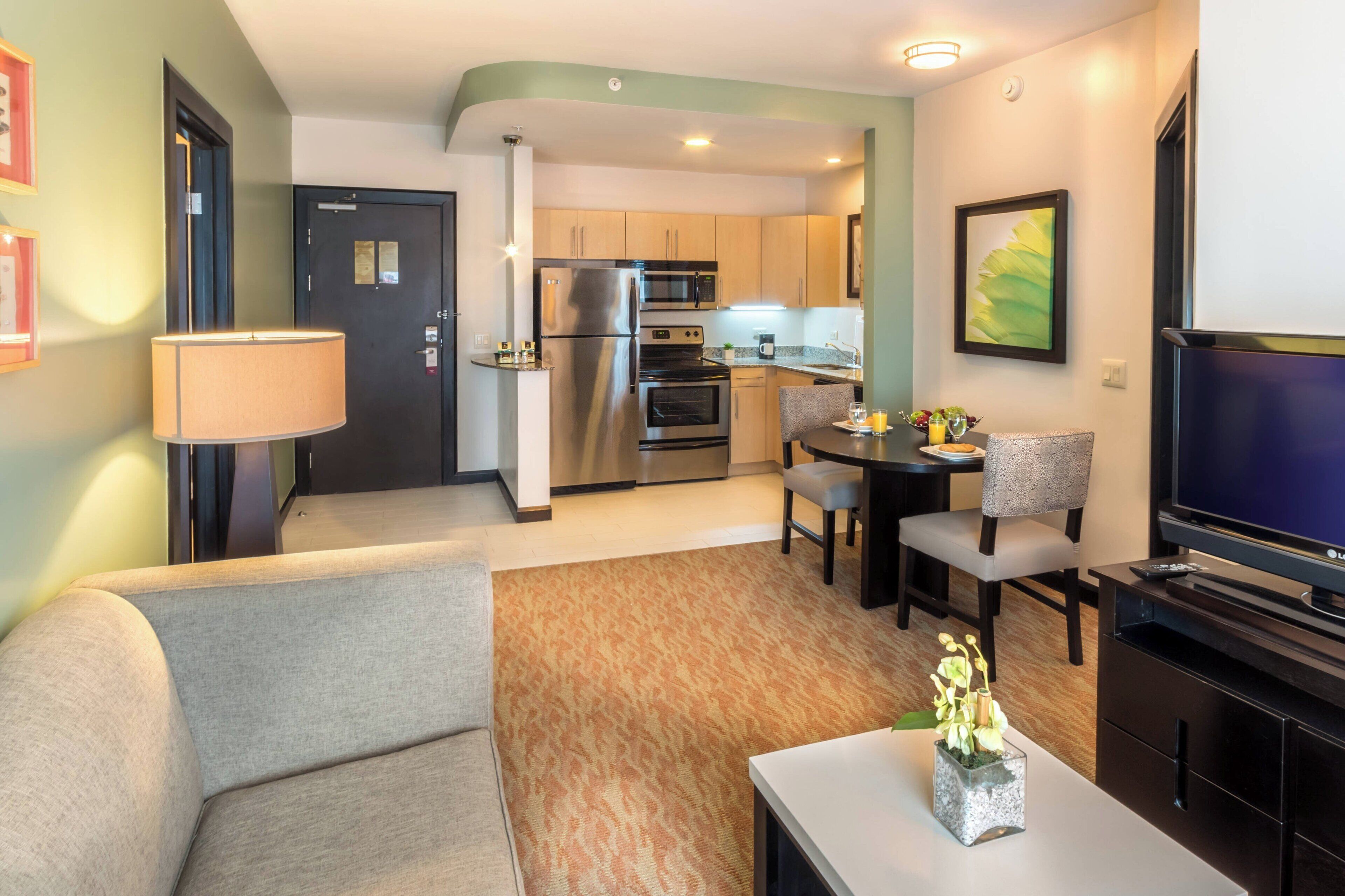 A compact living space with a sofa, a dining table with chairs, a TV, a kitchen area with a refrigerator and microwave, and a lamp.
