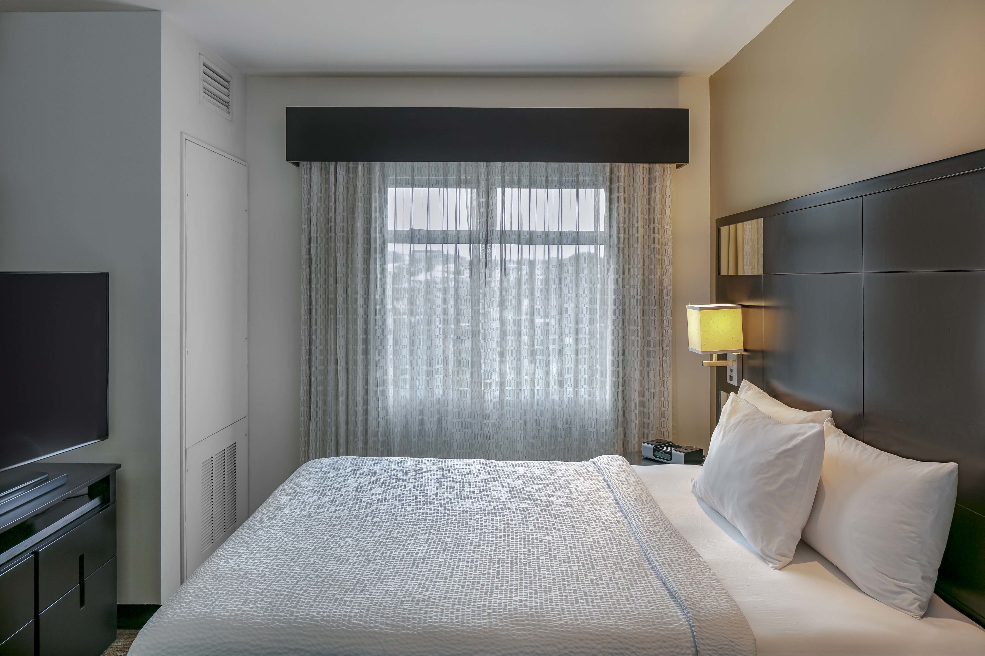 A hotel room with a large bed, a flat-screen TV, a bedside lamp, and a window with sheer curtains.