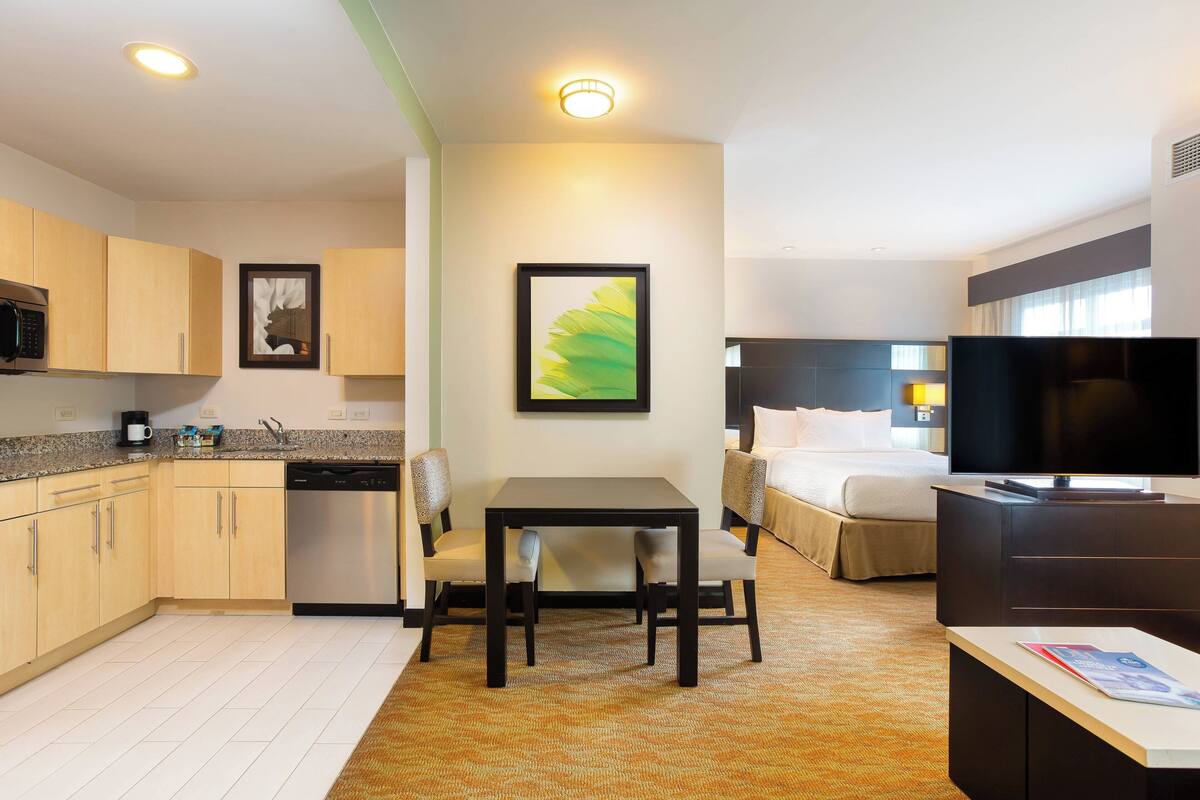 A compact hotel room with a kitchenette, a dining area, a bed, and a TV.