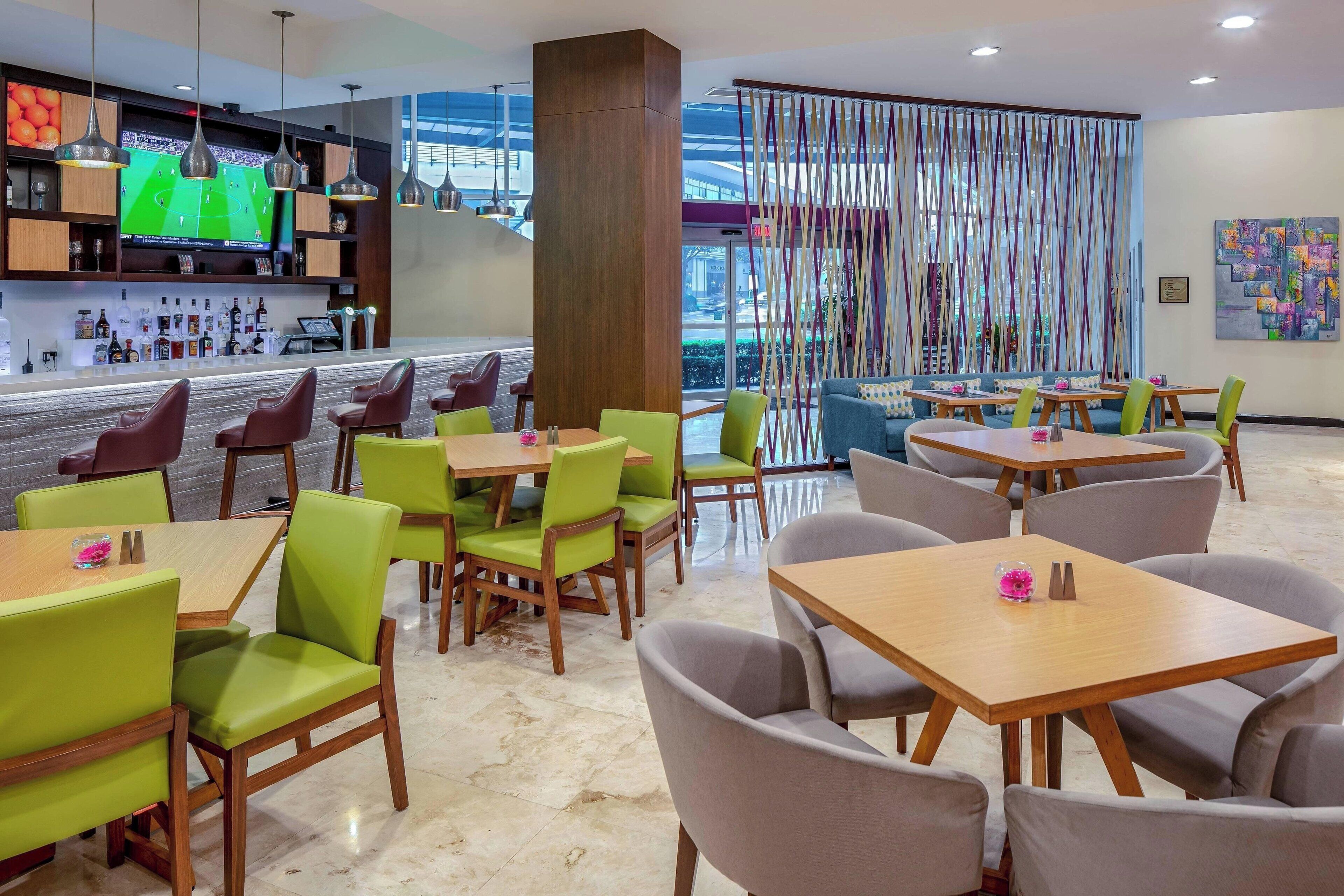 Een modern restaurant met groene en grijze stoelen, houten tafels, een bar met een televisie en uitzicht door grote ramen.