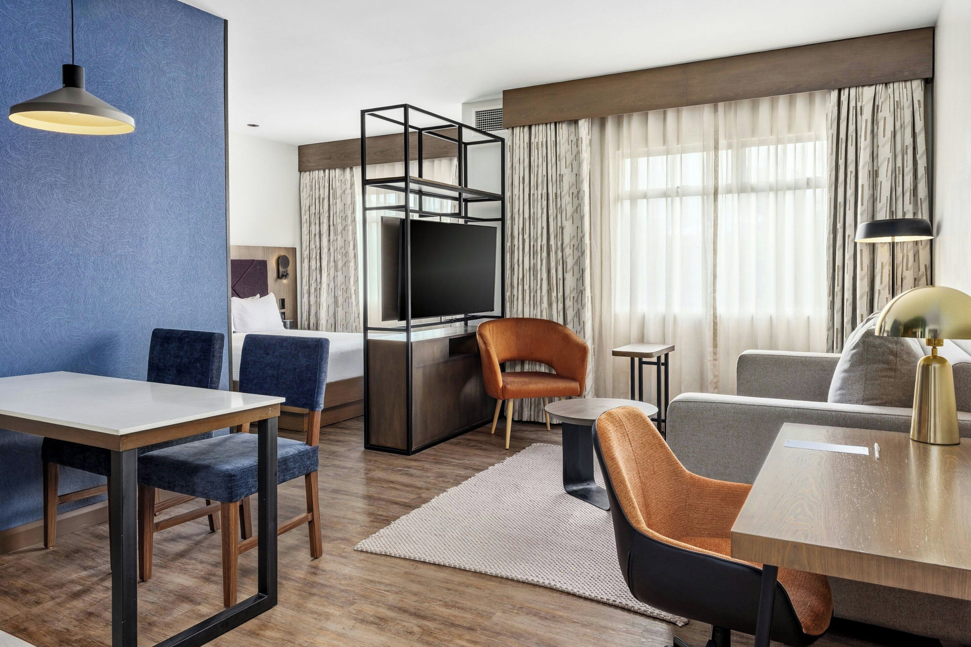 Ett modernt hotellrum med en matplats, en soffa, en TV och ett stort fönster med gardiner.
