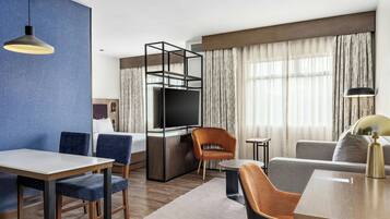 Ett modernt hotellrum med en matplats, en soffa, en TV och ett stort fönster med gardiner.