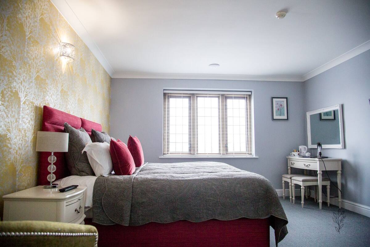 A bedroom with a bed, bedside table, lamp, dresser, and a window with blinds.