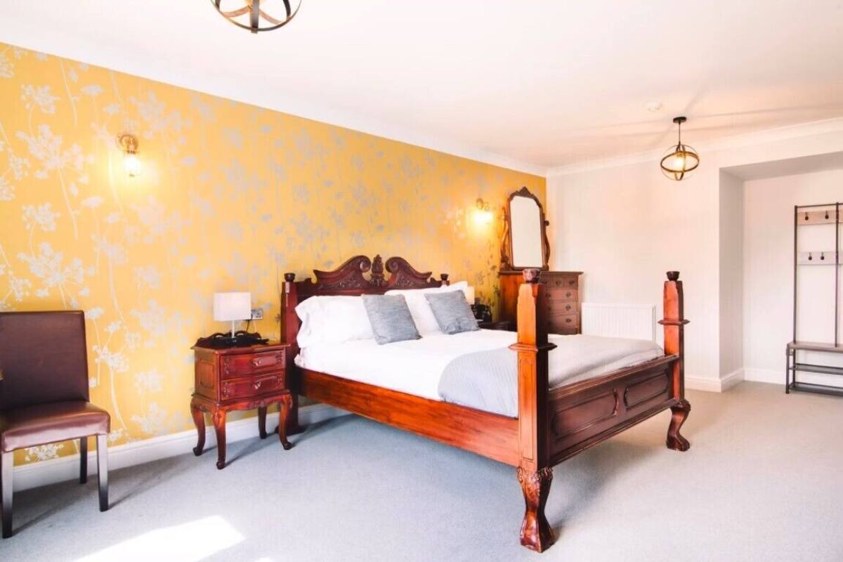A four-poster bed with a wooden frame, a nightstand with a lamp, and a chair in a room with yellow floral wallpaper.
