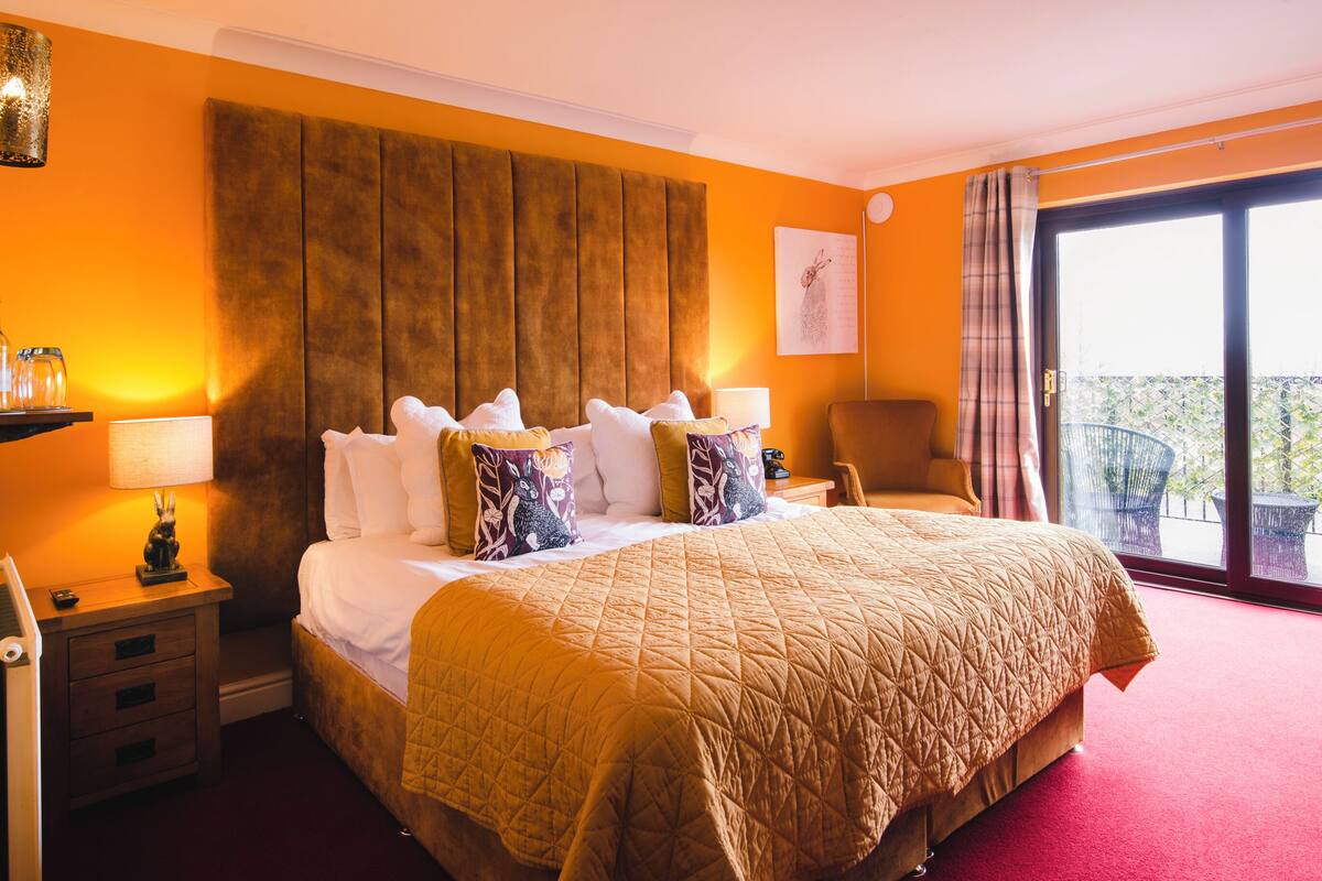 A hotel room with a large bed, a nightstand with a lamp, a chair, and a balcony view.